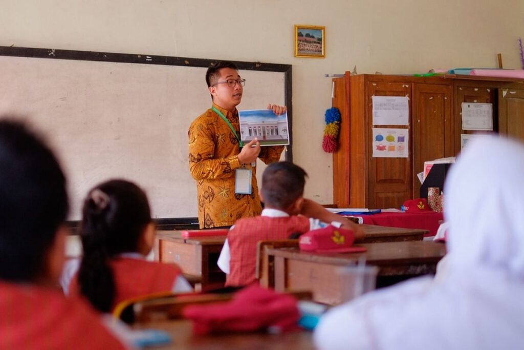 Seorang guru honorer sedang mengajar di kelas dengan antusias, menunjukkan materi pelajaran kepada siswa-siswi sekolah dasar di ruang kelas sederhana.